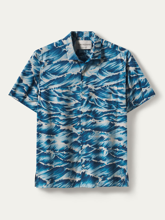 A short-sleeve button-up camp shirt with an all-over blue and white ocean wave pattern, displayed flat against a light beige background.
