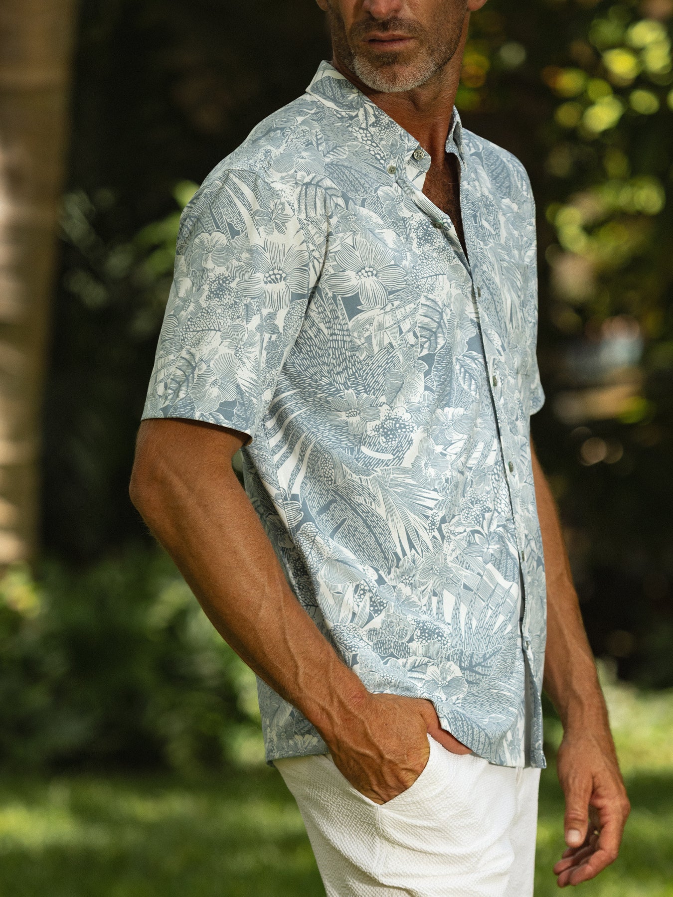 Green tropical-print short-sleeve shirt and white pants, front view outdoors, hand in pocket.