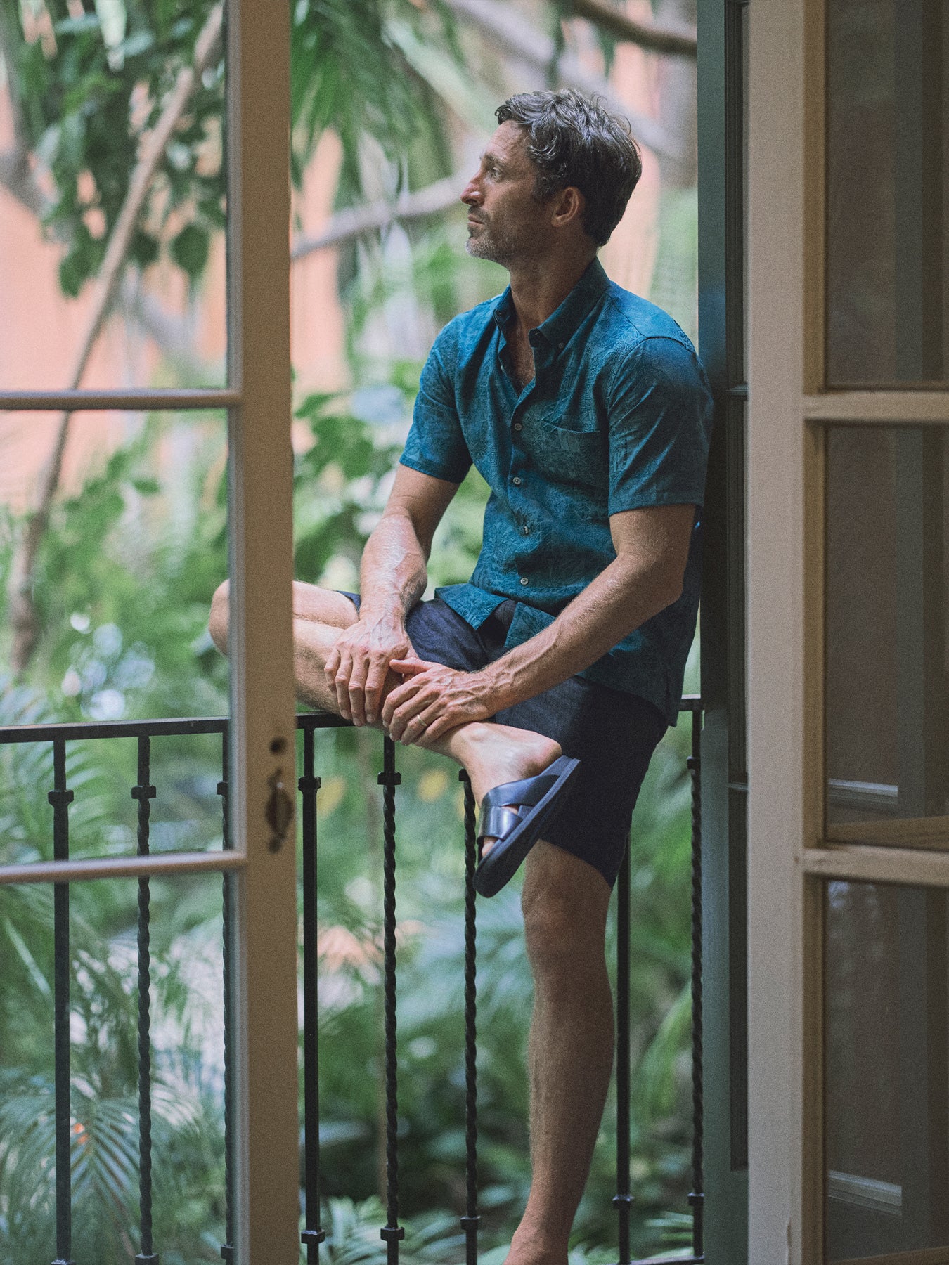 A man wearing a teal patterned short-sleeve button-up shirt and dark shorts sits cross-legged on a balcony railing, looking out over a lush green garden.