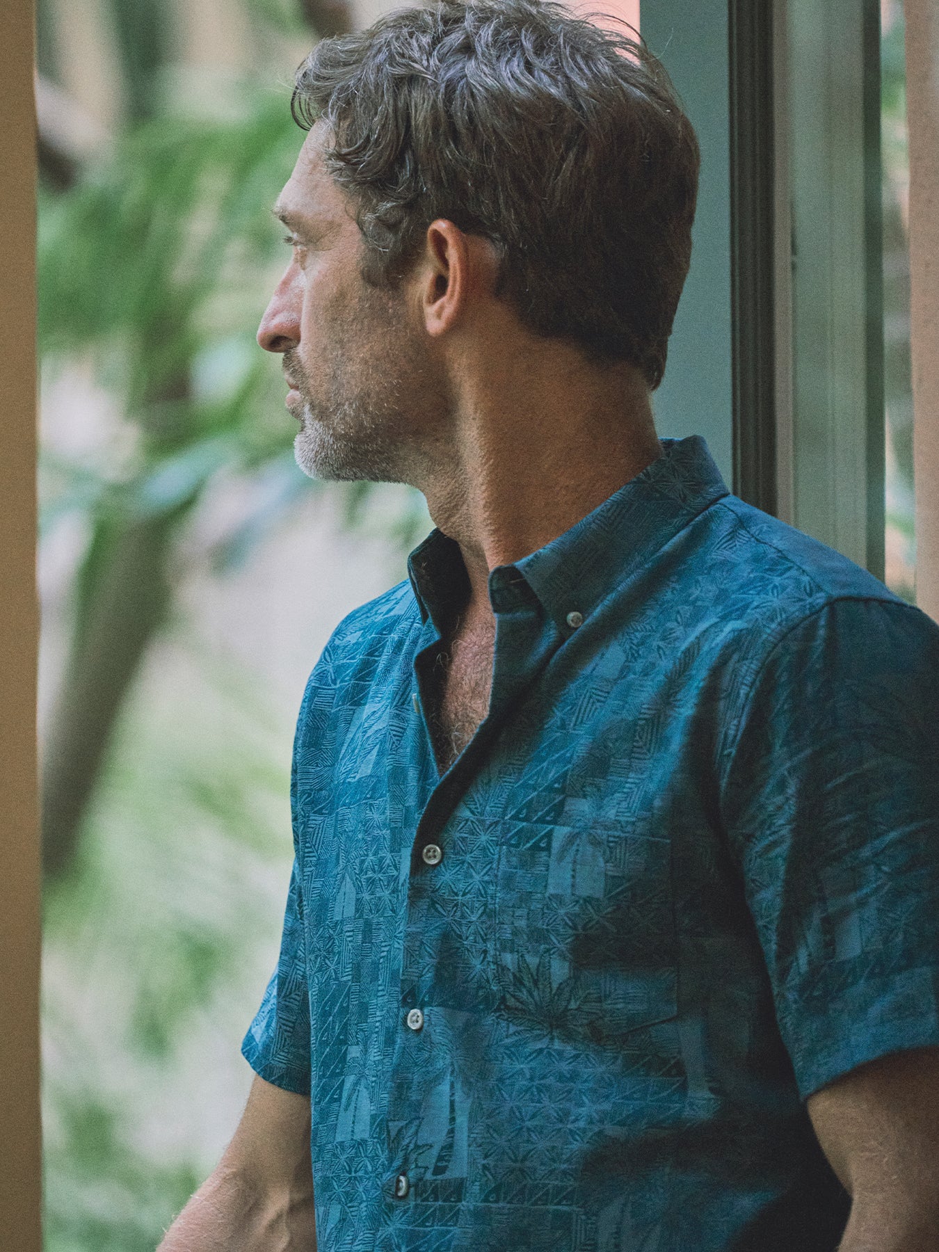A man in profile wearing a short-sleeve button-down shirt with a textured teal and blue tropical leaf pattern, standing near a window with blurred green foliage in the background.