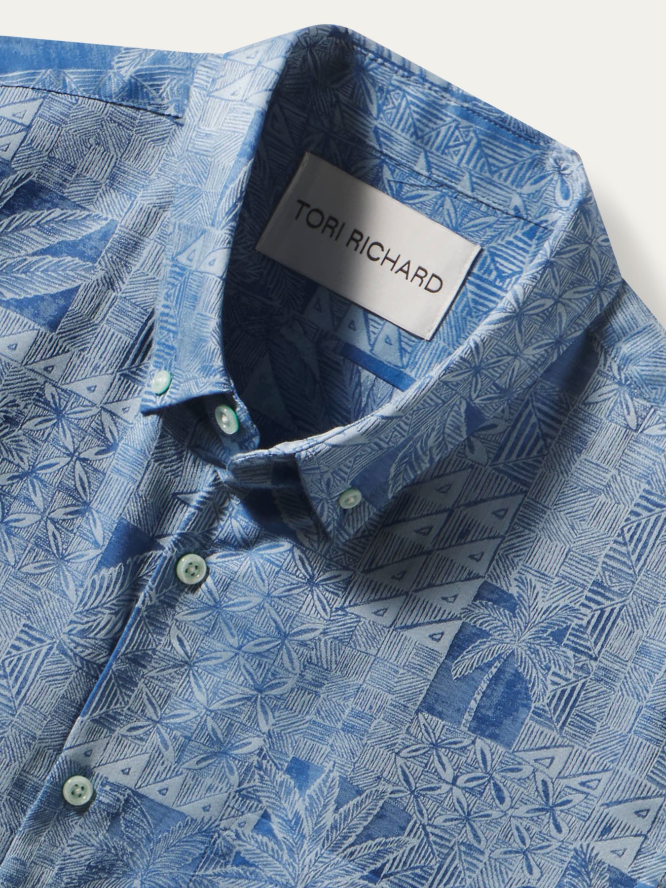 A close-up of a blue patterned button-down shirt by Tori Richard, featuring a geometric and botanical print in shades of blue and white, with a visible brand label on the collar.