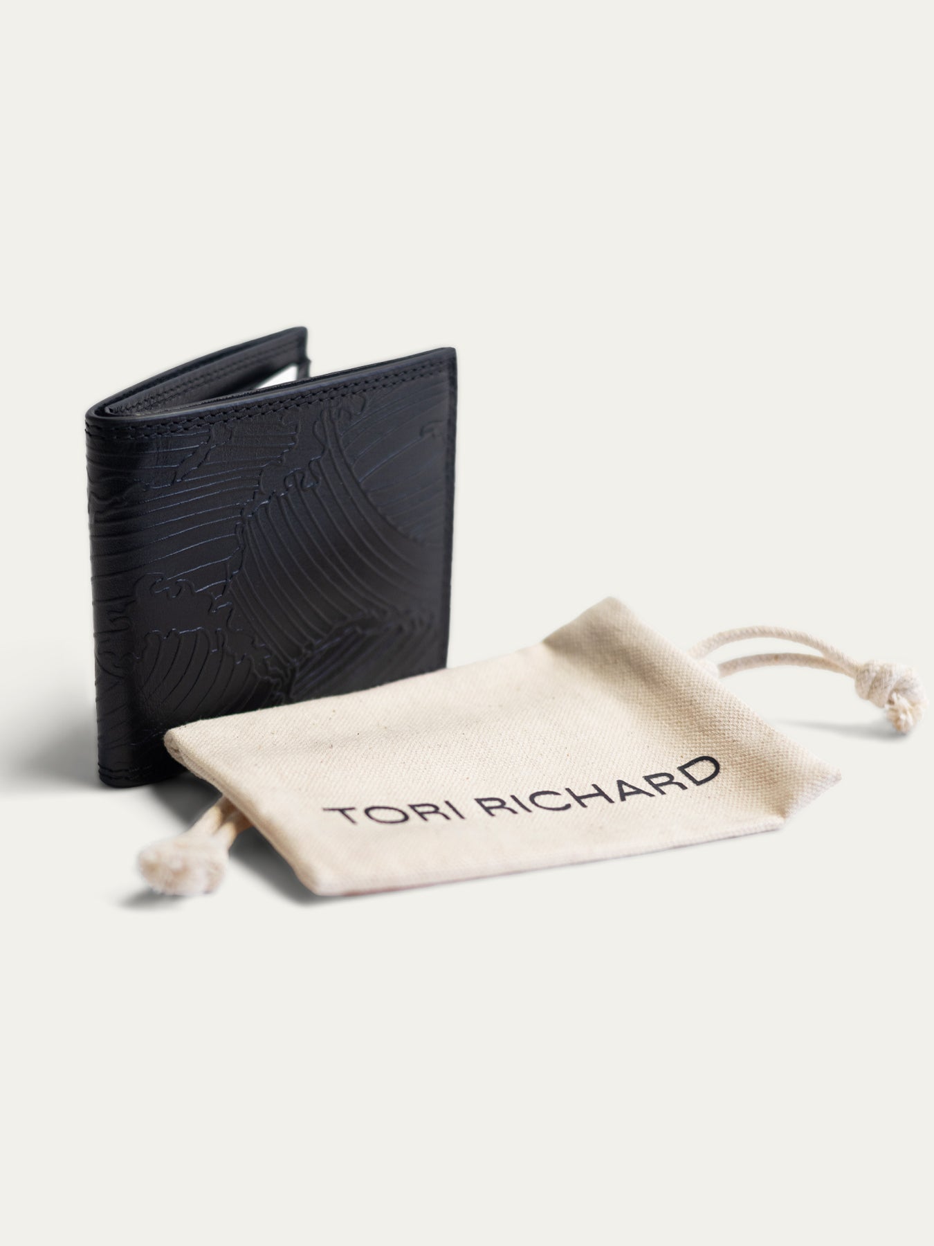 A black leather bifold wallet with an embossed wave pattern, shown next to a beige drawstring dust bag printed with the brand name 'TORI RICHARD' on a clean off-white background.