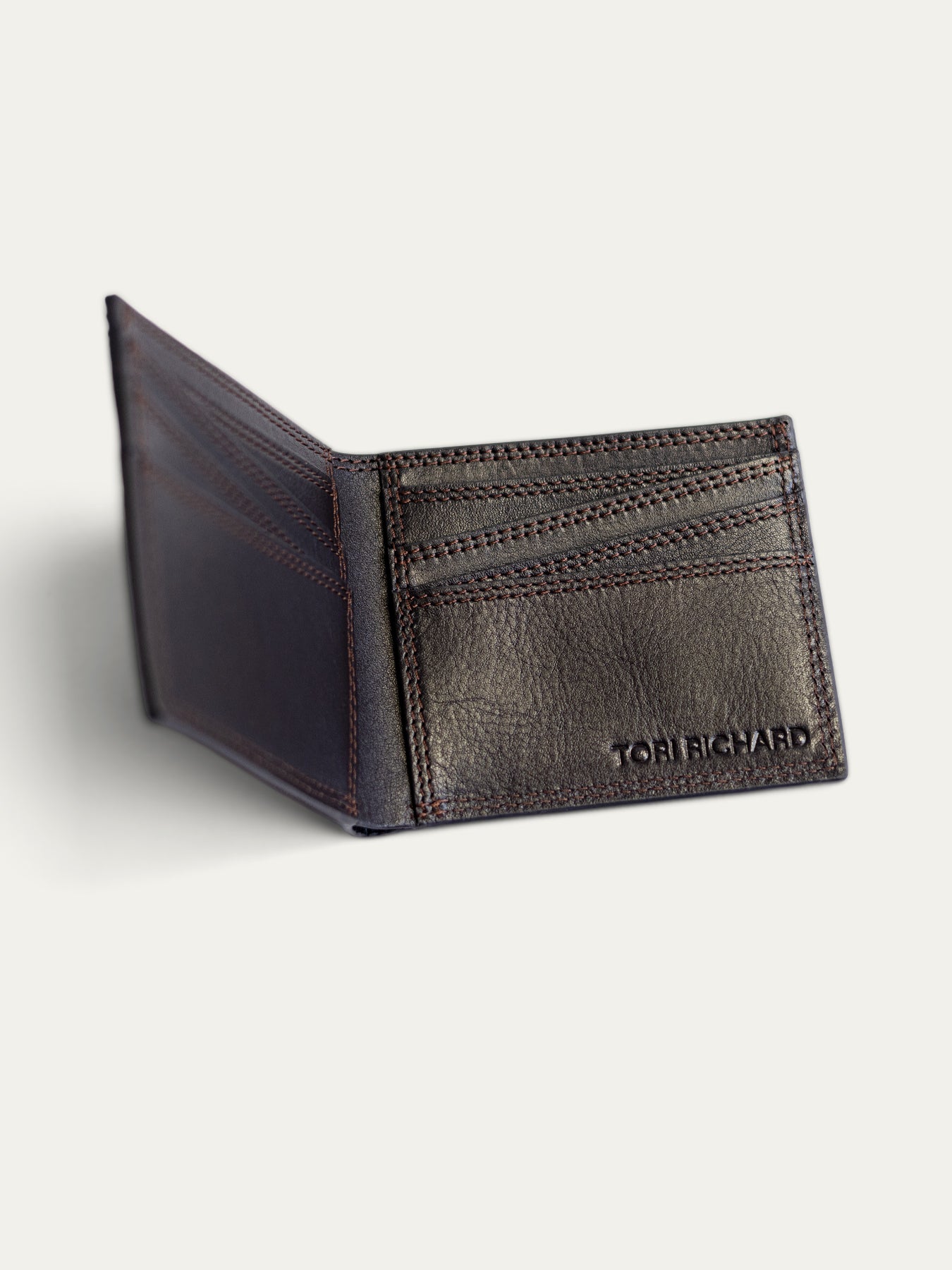 A dark brown leather bi-fold wallet shown open, featuring card slots on the front and the debossed brand name 'TORI RICHARD' on the lower right corner, set against a light off-white background.