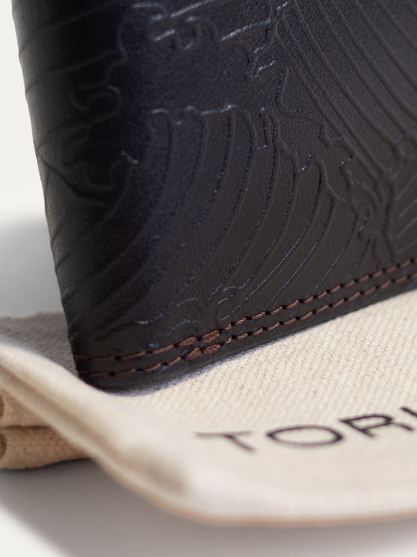 Close-up of a black leather wallet with embossed wave-like detailing and contrast stitching, resting on a beige branded dust bag with partial 'TORI' text visible.