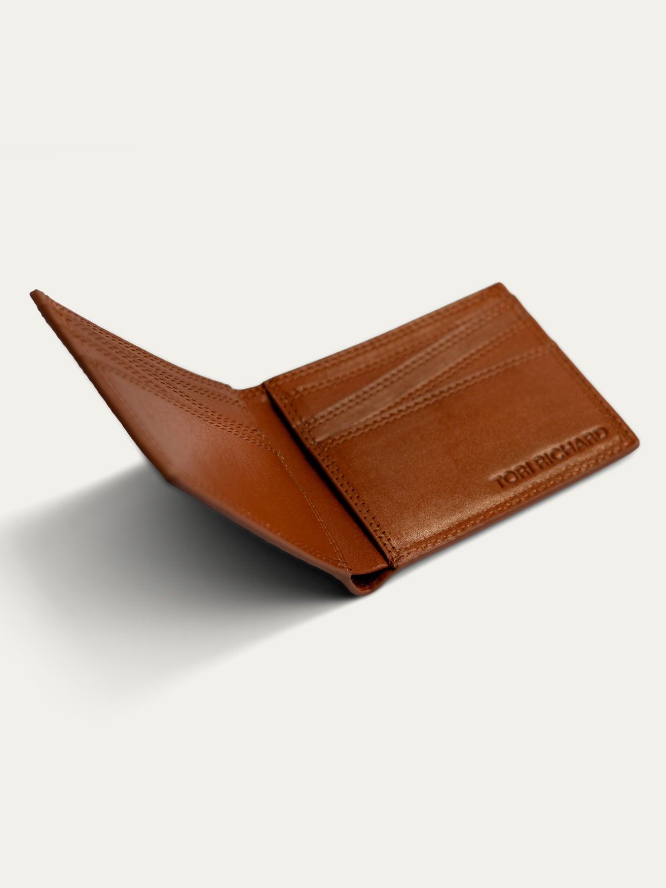 A brown leather bifold wallet shown open, displaying its interior card slots and a debossed 'TORI Richard' logo on the right side, set against a light off-white background with a soft shadow.