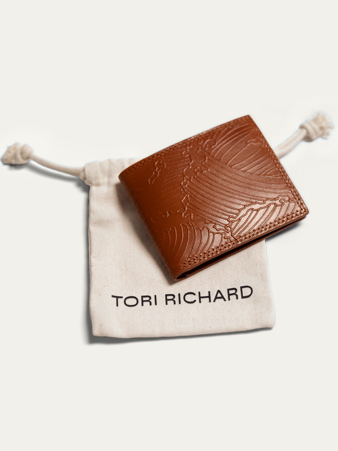 A brown leather bifold wallet with an embossed wave pattern, resting on a beige drawstring pouch printed with the brand name TORI RICHARD.