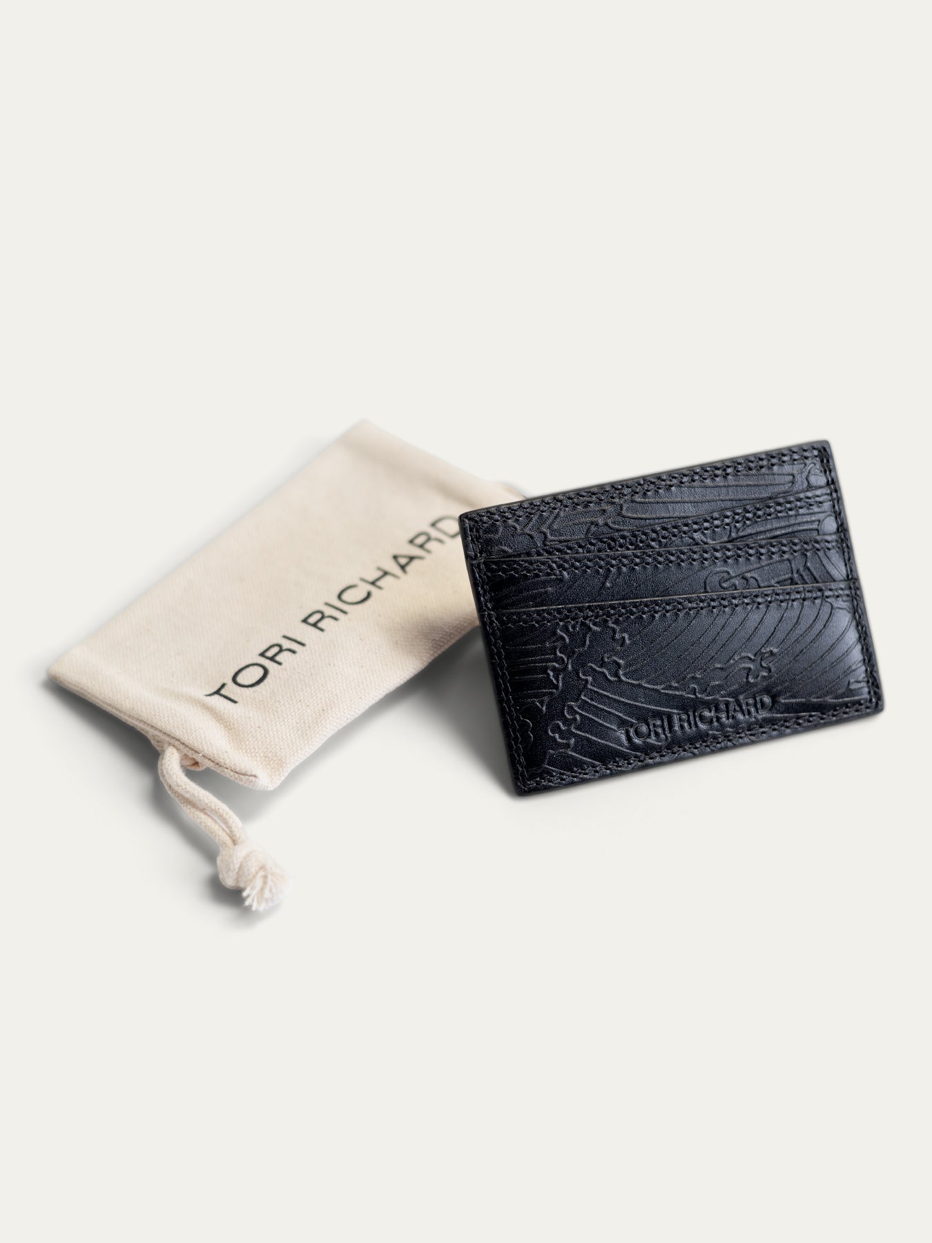 A black textured leather card holder by Tori Richardson, displayed next to its beige branded dust bag on a light cream background.