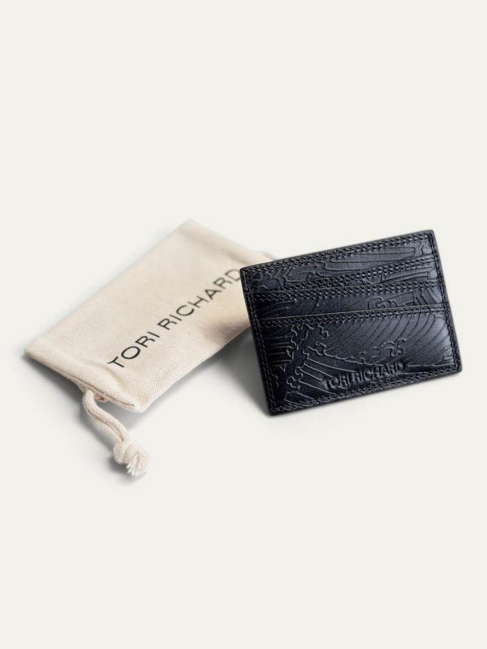A black textured leather card holder by Tori Richardson, displayed next to its beige branded dust bag on a light cream background.