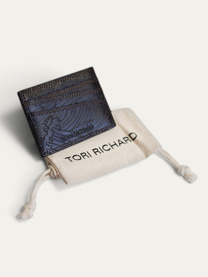 A dark navy blue textured leather card holder wallet with an embossed winged design, shown with its cream-colored drawstring dust bag printed with the brand name 'TORI RICHARD'.