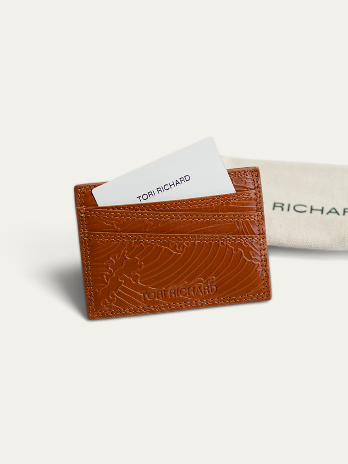 A brown embossed leather card holder wallet by Tori Richard, shown with a white card partially inserted and a beige branded dust bag behind it on a light neutral background.