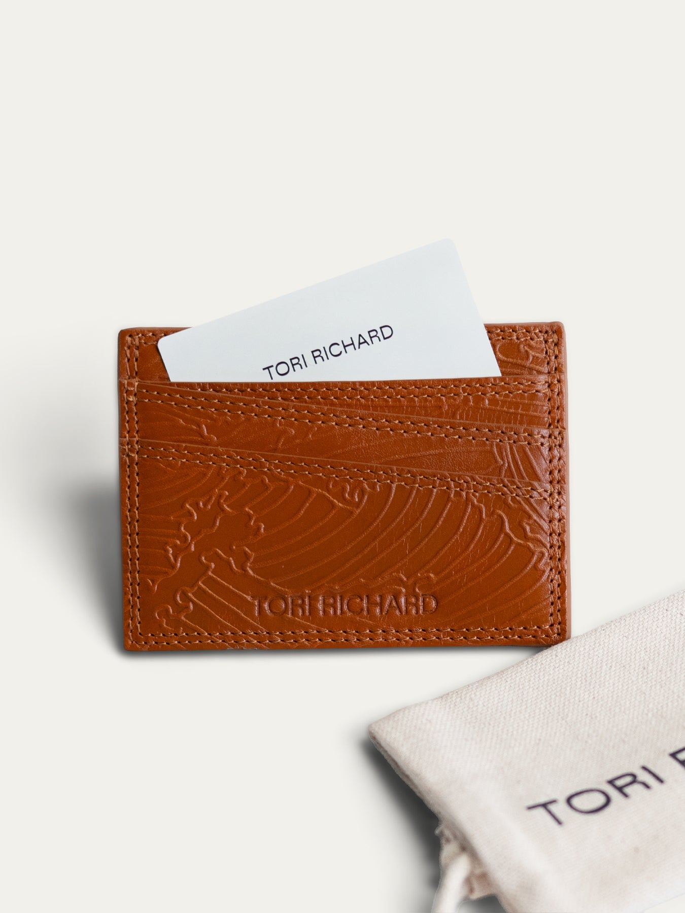 A brown embossed leather card holder with a wave pattern, shown with a white card and a branded dust bag on a light beige background.
