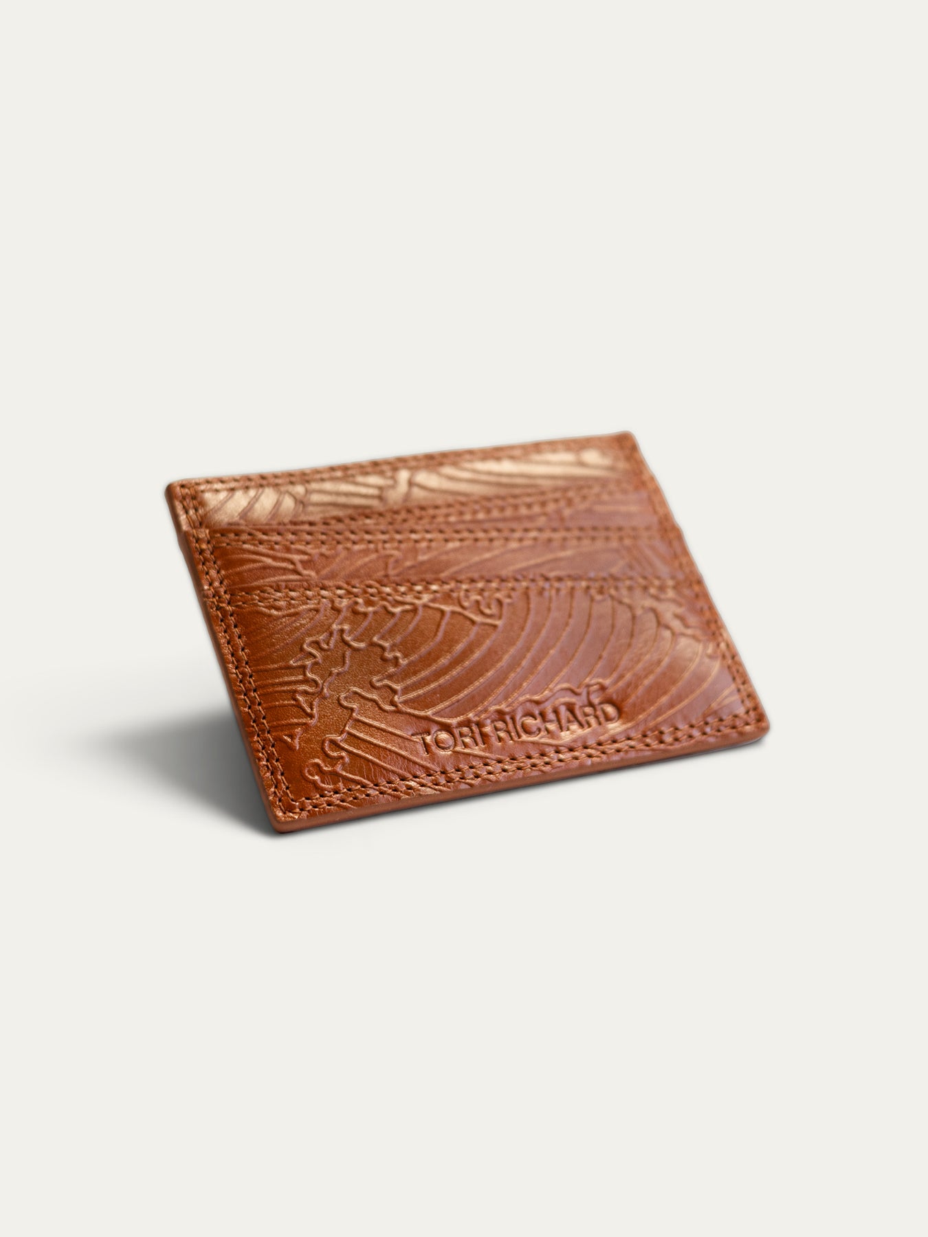A tan leather card holder wallet by Josh Richard, featuring an embossed wave pattern and brand name, shown on a light gray background.
