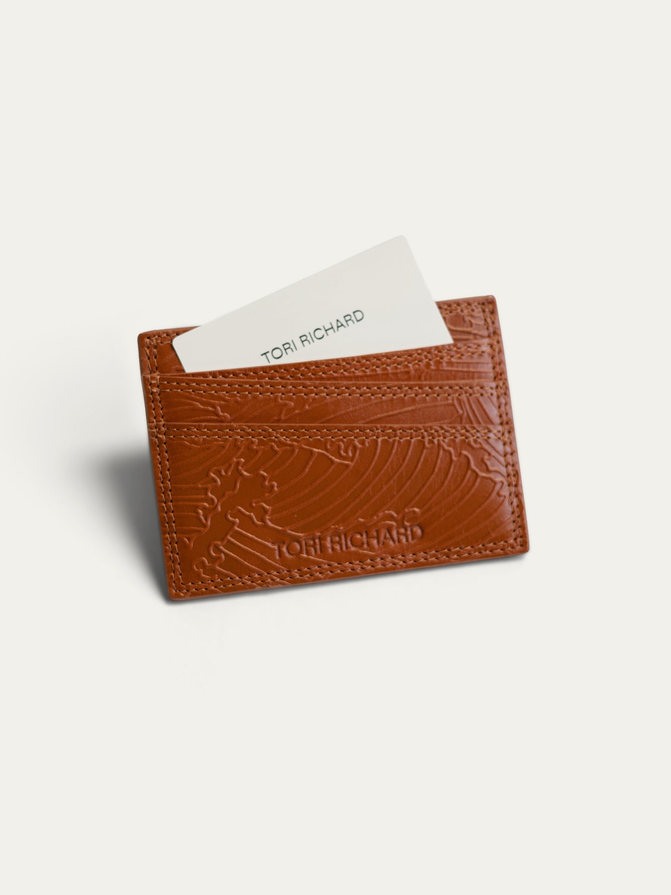 A brown embossed leather card holder wallet by Tori Richard, shown with a white card partially inserted, on a light off-white background.