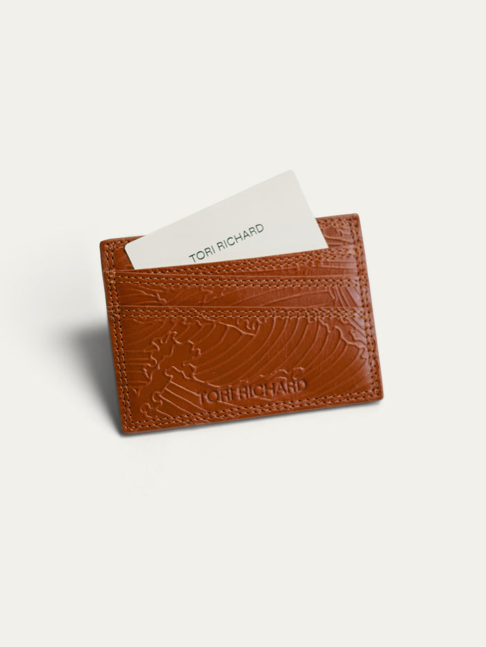 A brown embossed leather card holder wallet by Tori Richard, shown with a white card partially inserted, on a light off-white background.