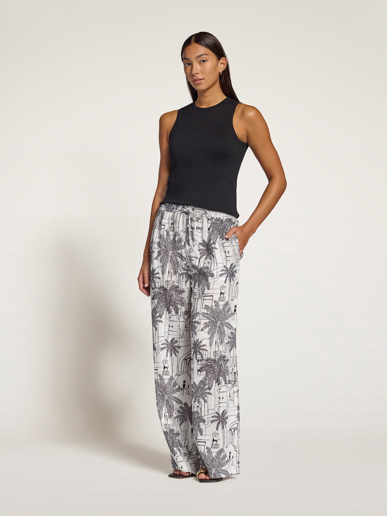A woman modeling wide-leg pants with a gray and white palm tree pattern, paired with a black sleeveless top, against a plain light gray studio background.