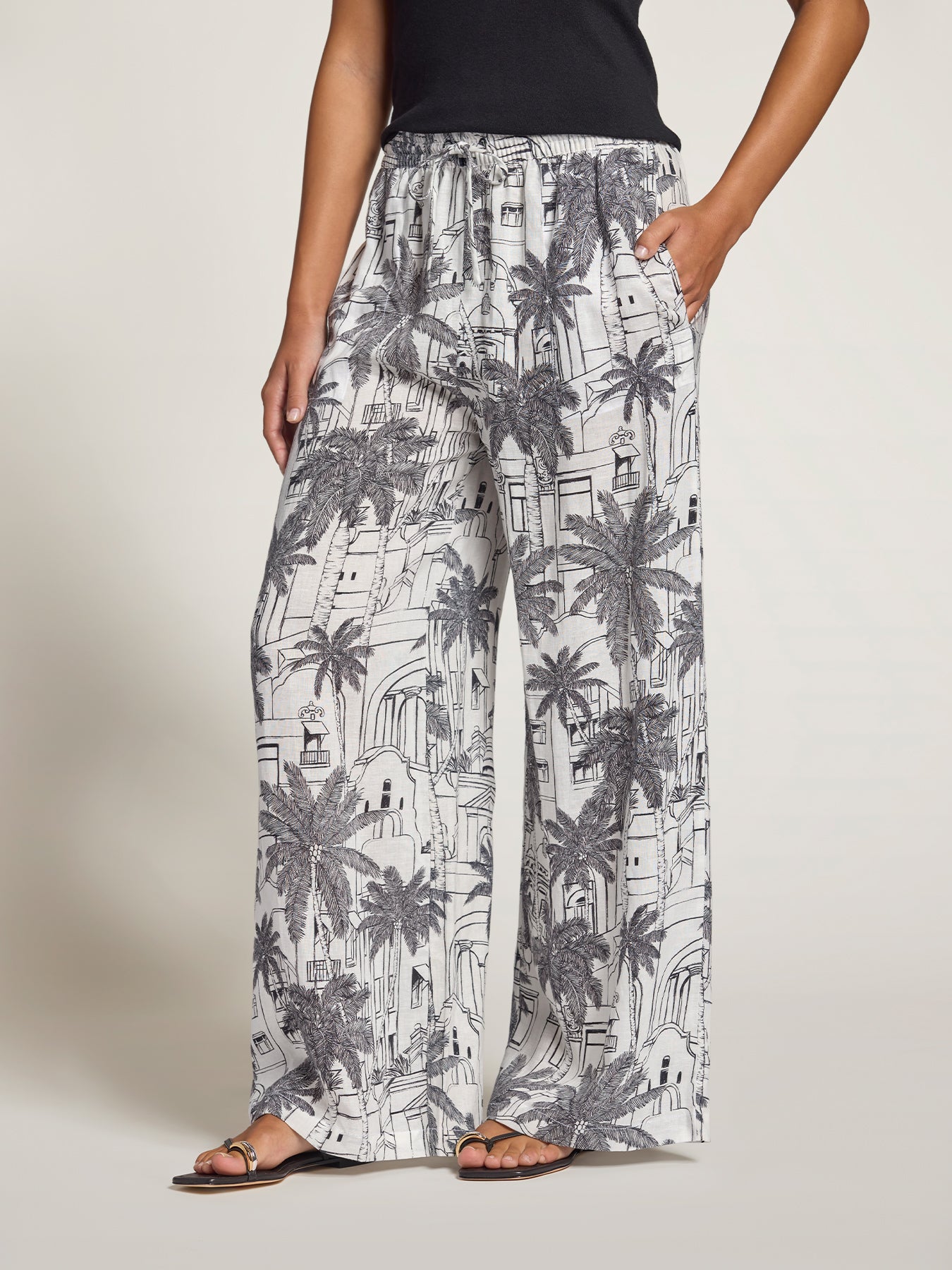 A person wearing loose, wide-leg pants with an allover black palm tree and architectural line drawing print on a white base, paired with a black top and flat sandals against a plain light beige studio background.