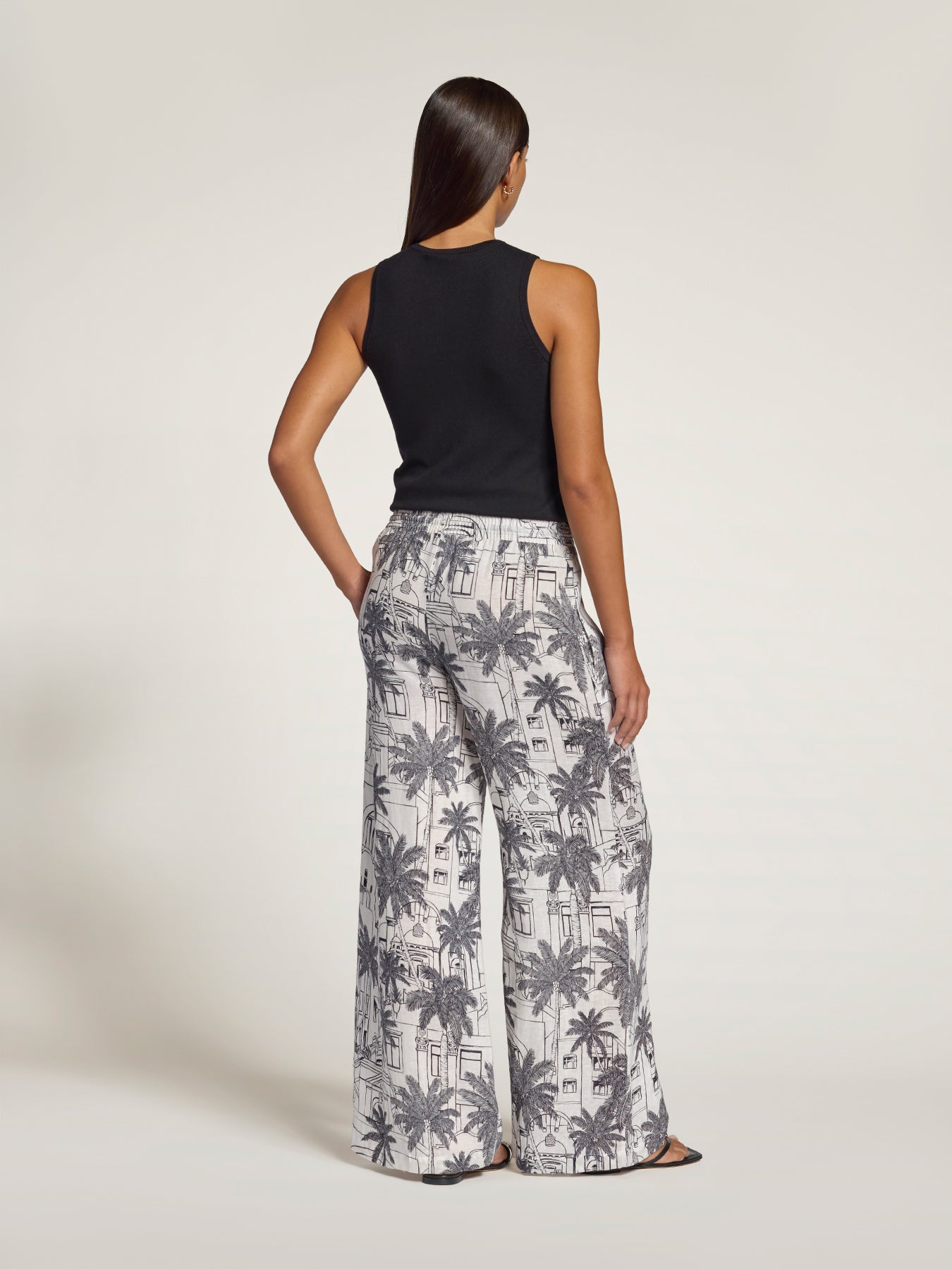 A woman shown from the back wearing a black sleeveless top and wide-leg pants with a gray and white palm tree print, photographed against a plain light studio background.