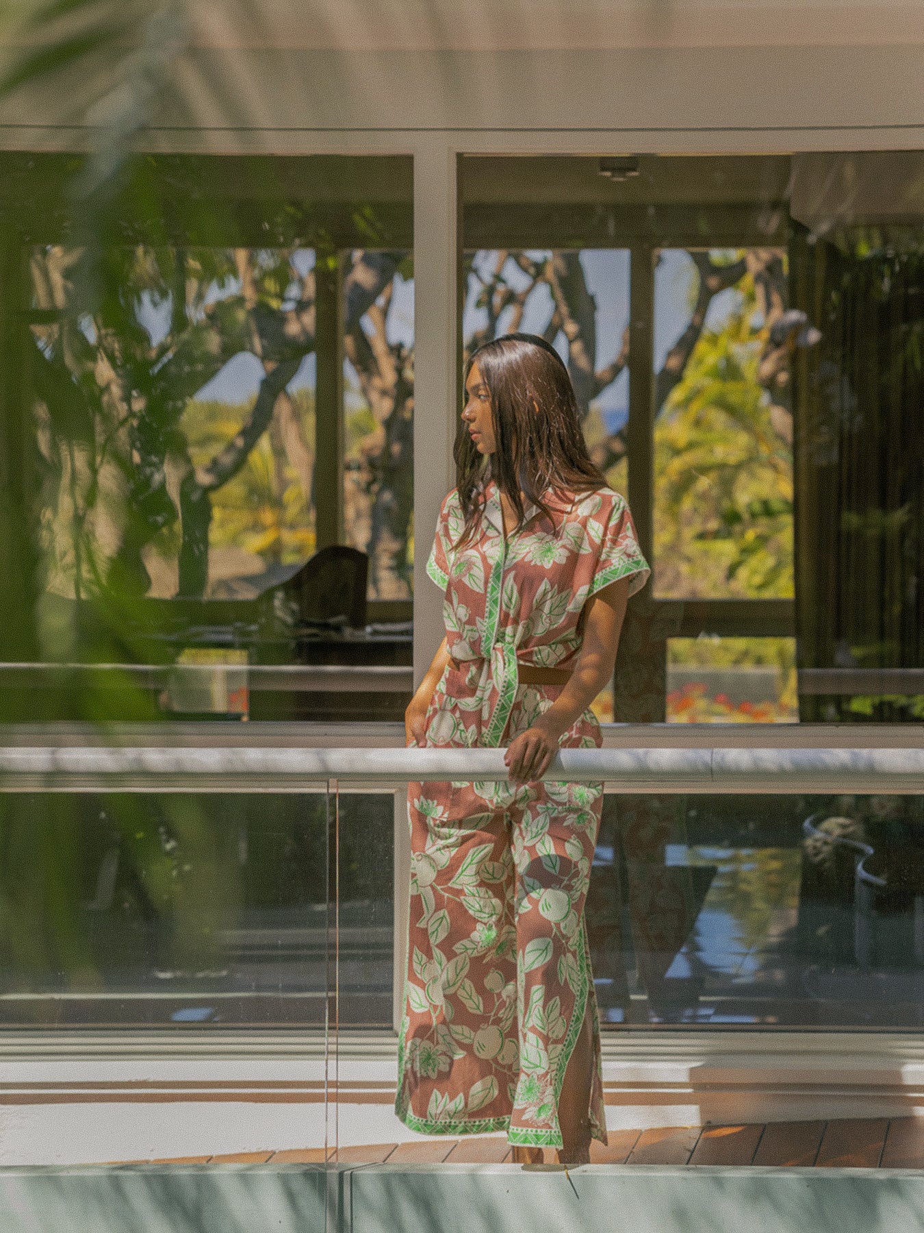 A woman wearing a matching short-sleeve top and maxi skirt set with a green and brown botanical pattern stands on a modern glass balcony overlooking a tropical garden.
