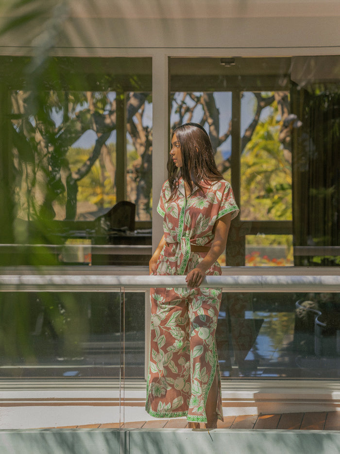 A woman wearing a matching short-sleeve top and maxi skirt set with a green and brown botanical pattern stands on a modern glass balcony overlooking a tropical garden.