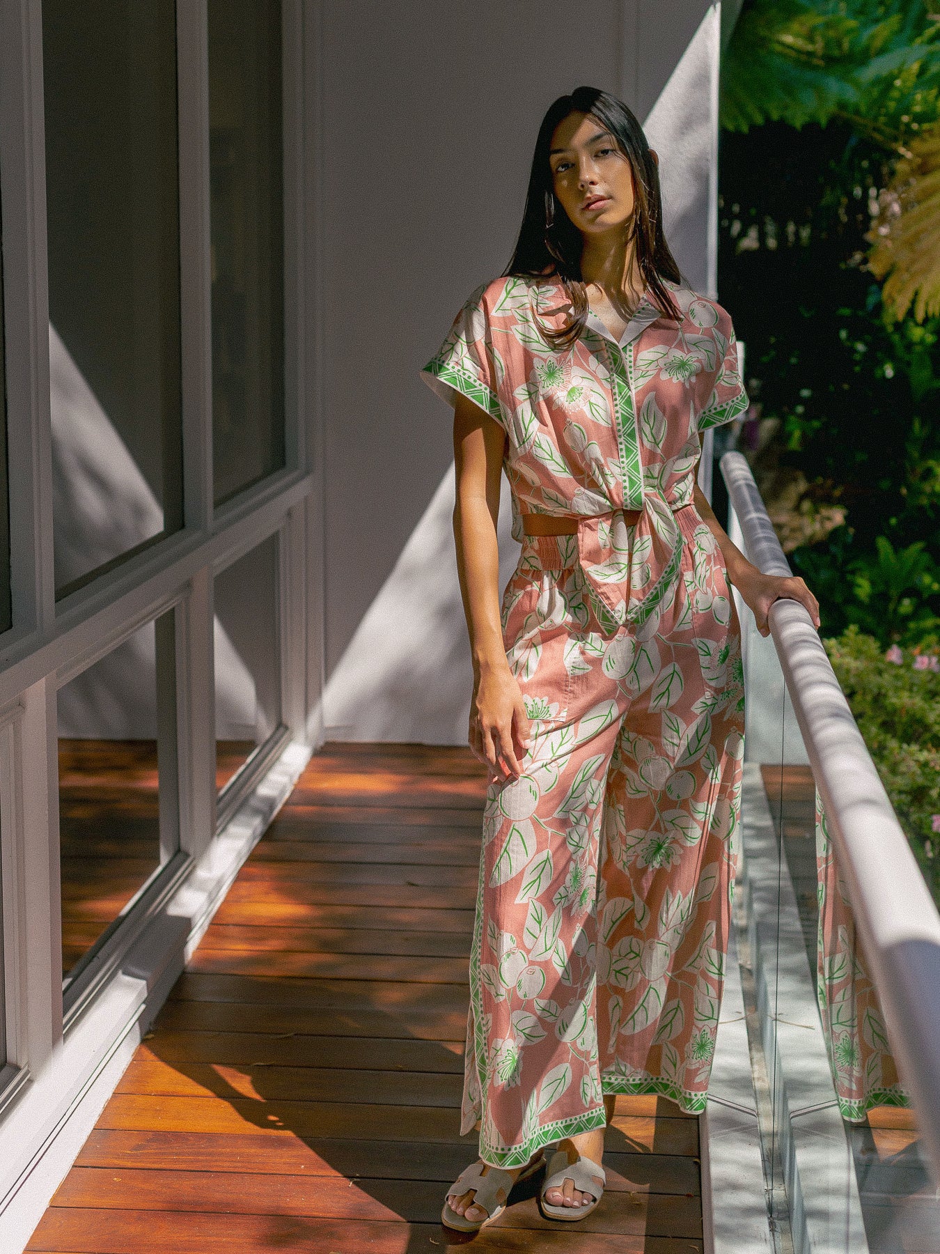 A woman stands on a wooden deck balcony wearing a pink and green tropical leaf print maxi dress with a tie waist and short sleeves.