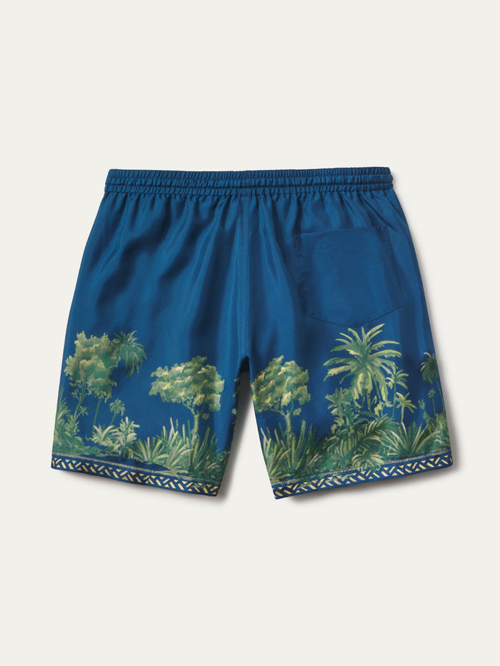 Blue swim trunks with a green tropical palm tree print and a geometric pattern border, shown flat on a light beige background.
