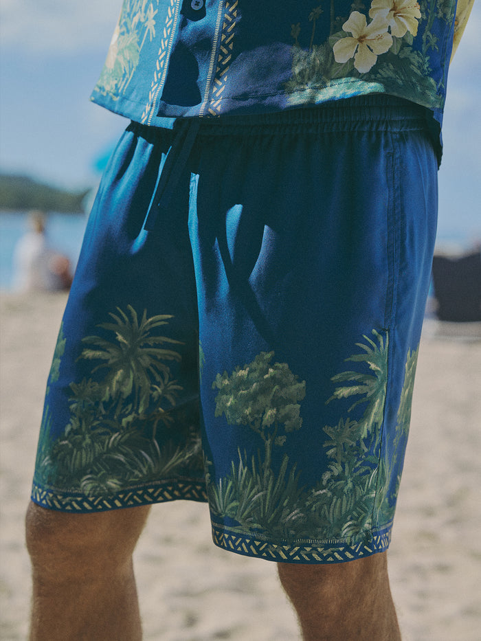 A close-up of a person wearing blue tropical print swim shorts and a matching blue shirt on a sandy beach.
