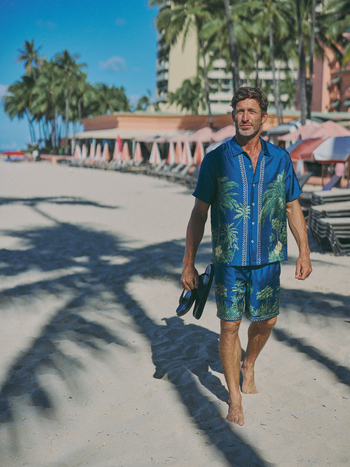 A man walks barefoot on a sandy beach wearing a blue tropical print short-sleeve button-up shirt and matching blue shorts, holding a pair of black sandals. Palm trees, beach umbrellas, and buildings are visible in the background under a blue sky.