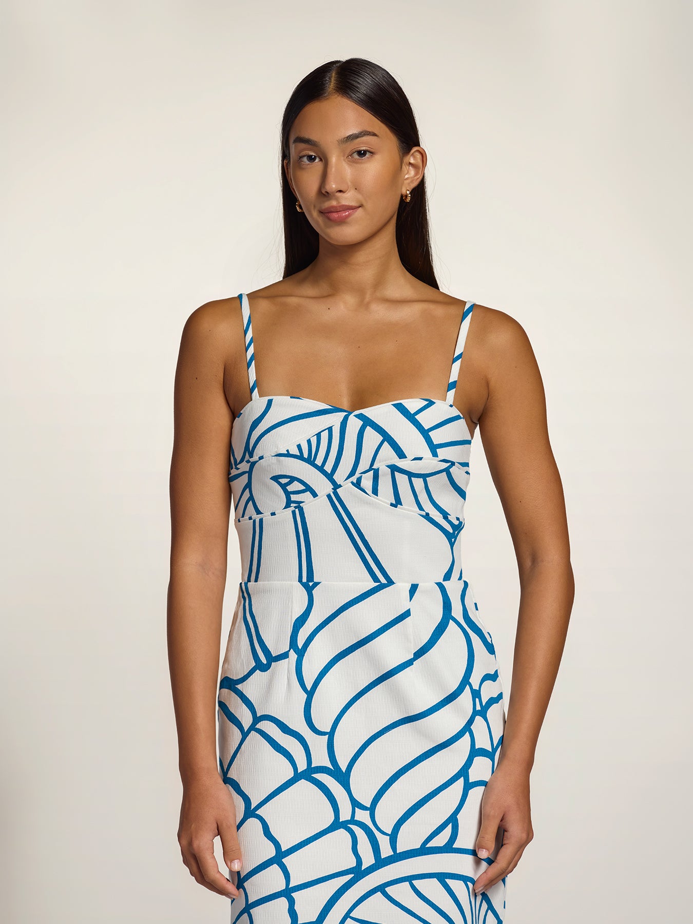 A woman modeling a white spaghetti-strap dress with a bold blue abstract line pattern, set against a plain light beige studio background.