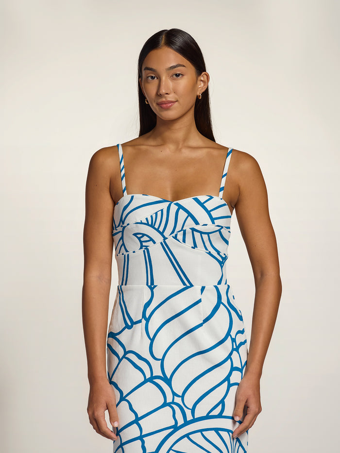 A woman modeling a white spaghetti-strap dress with a bold blue abstract line pattern, set against a plain light beige studio background.