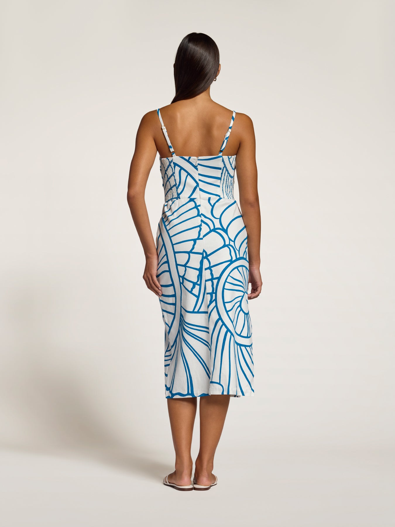 A woman viewed from the back wearing a sleeveless midi slip dress with thin straps and a blue abstract line pattern on a white base, standing against a plain light beige studio background.