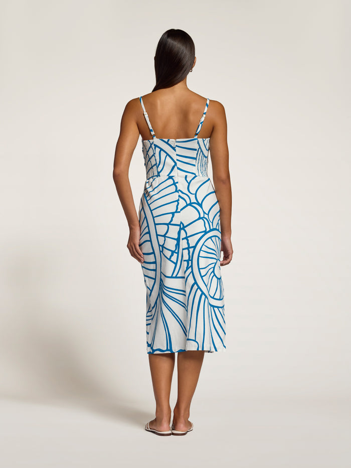 A woman viewed from the back wearing a sleeveless midi slip dress with thin straps and a blue abstract line pattern on a white base, standing against a plain light beige studio background.