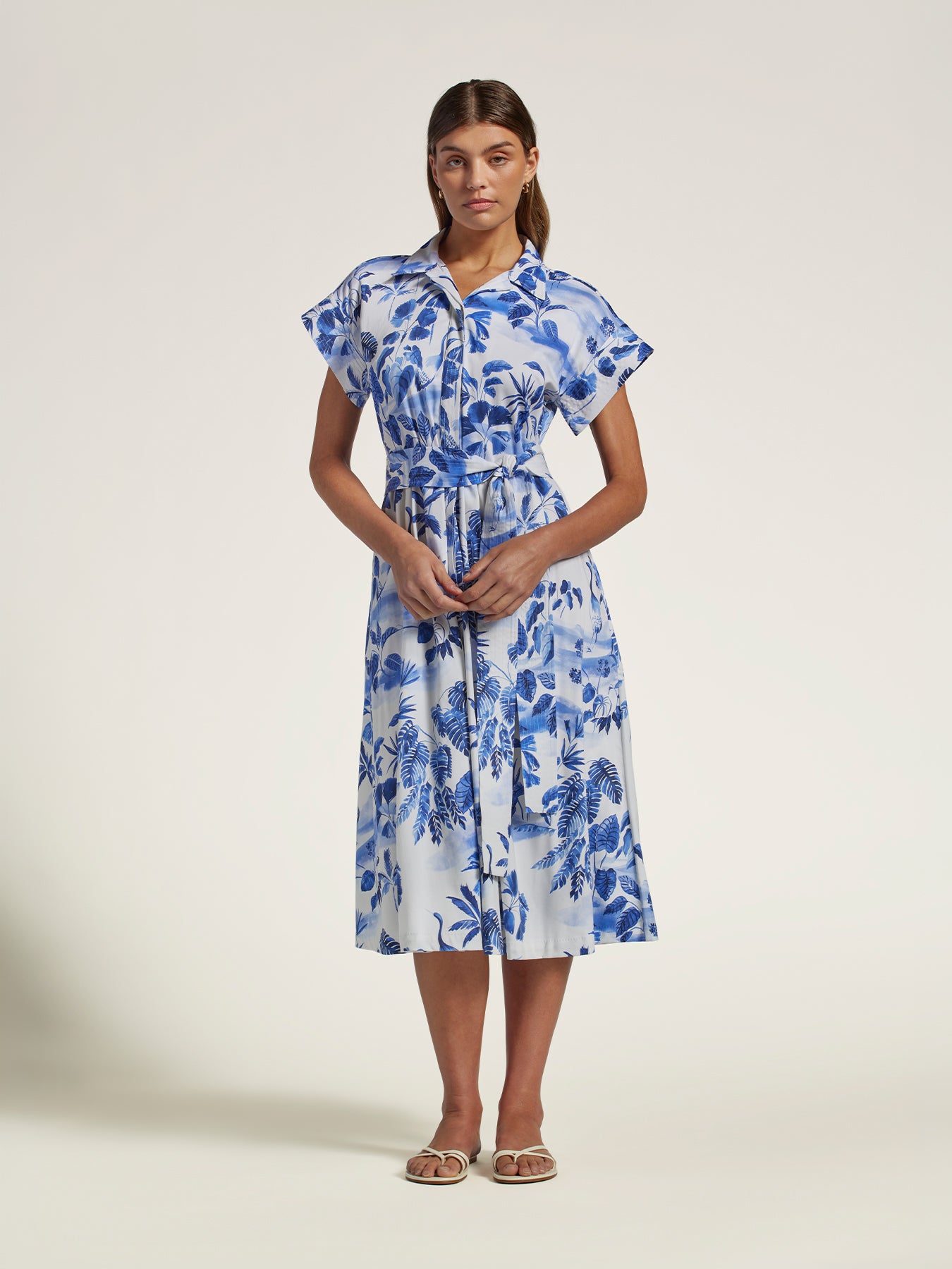 A woman modeling a white midi shirt dress with a blue tropical floral print, featuring ruffled short sleeves, a button-up front, and a matching fabric tie belt at the waist, set against a plain light beige studio background.