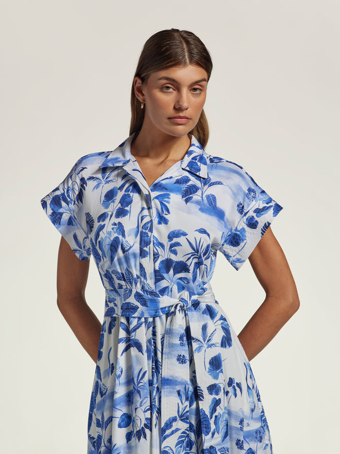 A woman modeling a short-sleeve collared dress with a blue and white tropical floral print, set against a plain light beige studio background.