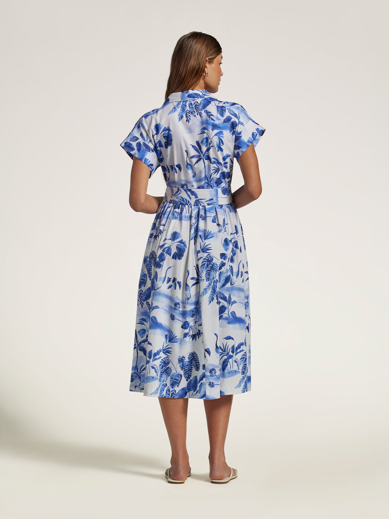 A woman viewed from the back wearing a blue and white floral print midi dress with short flutter sleeves and a tie waist, standing against a plain light beige studio background.