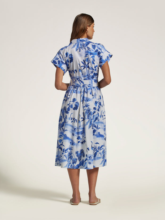 A woman viewed from the back wearing a blue and white floral print midi dress with short flutter sleeves and a tie waist, standing against a plain light beige studio background.