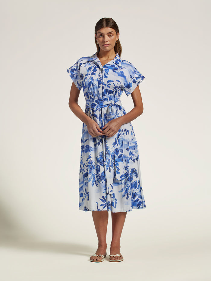 A woman modeling a white midi shirt dress with a blue tropical floral print, featuring ruffled short sleeves, a button-up front, and a matching fabric tie belt at the waist, set against a plain light beige studio background.