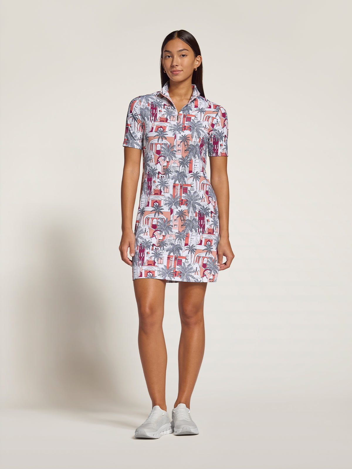 A woman modeling a short-sleeve collared mini dress with a colorful tropical and architectural print, paired with white sneakers, against a plain light beige studio background.
