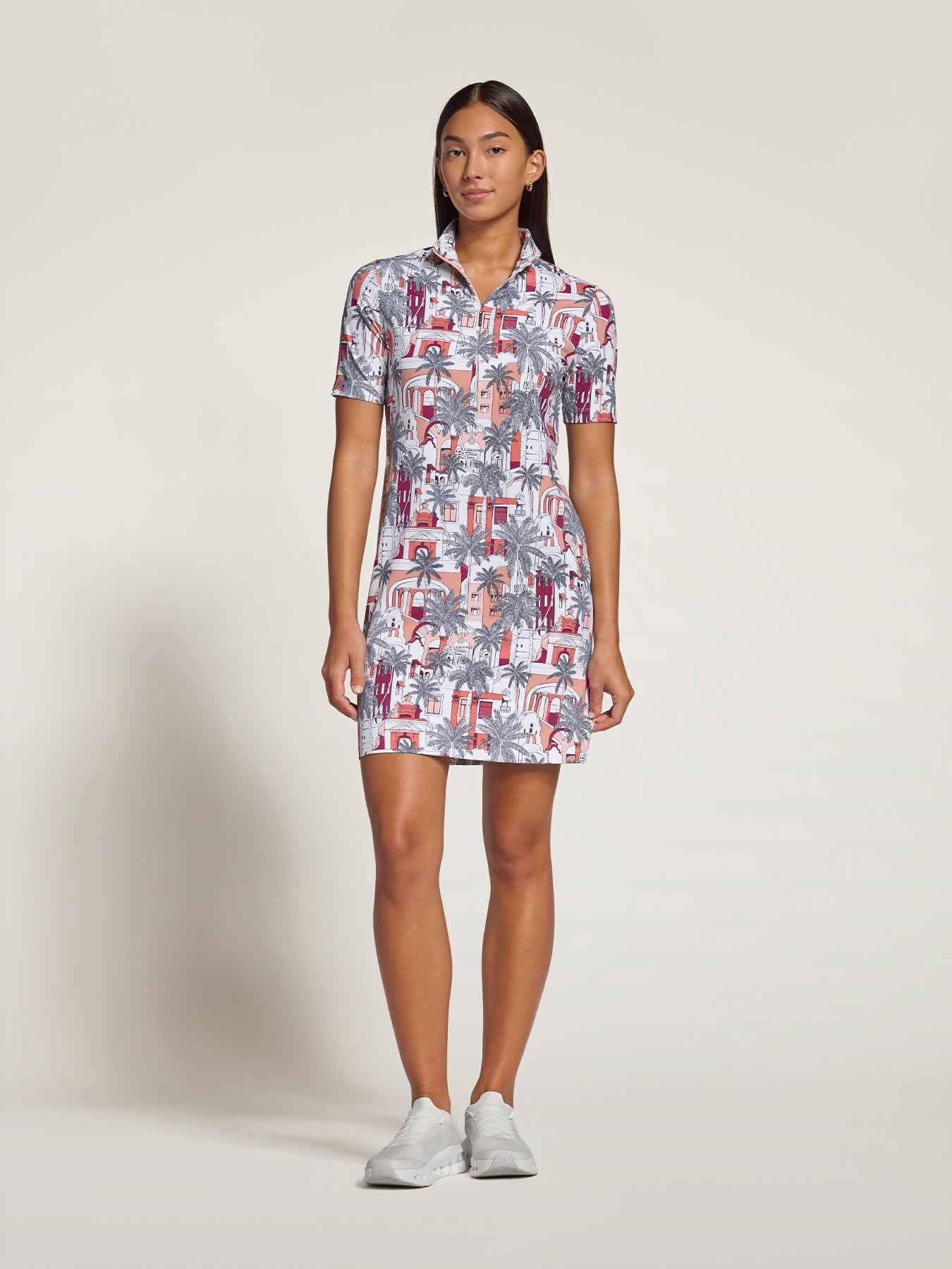 A woman modeling a short-sleeve collared mini dress with a colorful tropical and architectural print, paired with white sneakers, against a plain light beige studio background.