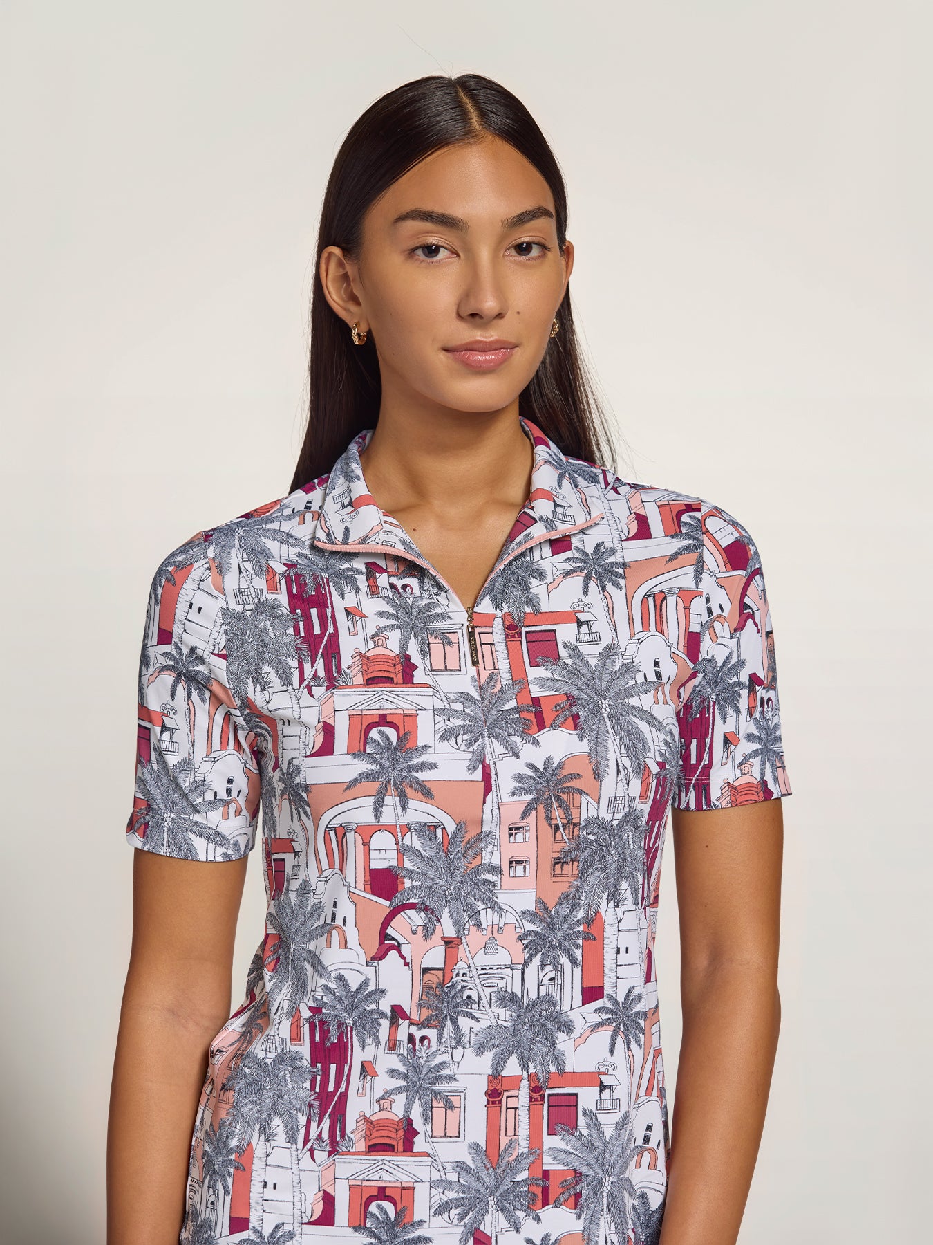 A woman modeling a short-sleeve collared button-up shirt with an all-over architectural and palm tree print in coral, gray, and burgundy on a white base, photographed against a plain light beige studio background.