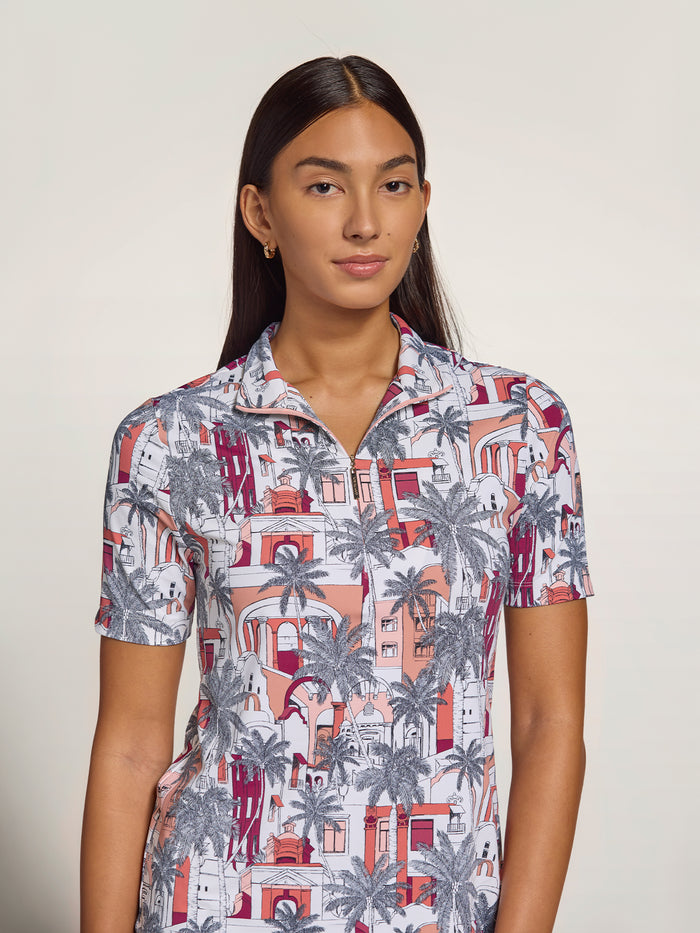 A woman modeling a short-sleeve collared button-up shirt with an all-over architectural and palm tree print in coral, gray, and burgundy on a white base, photographed against a plain light beige studio background.