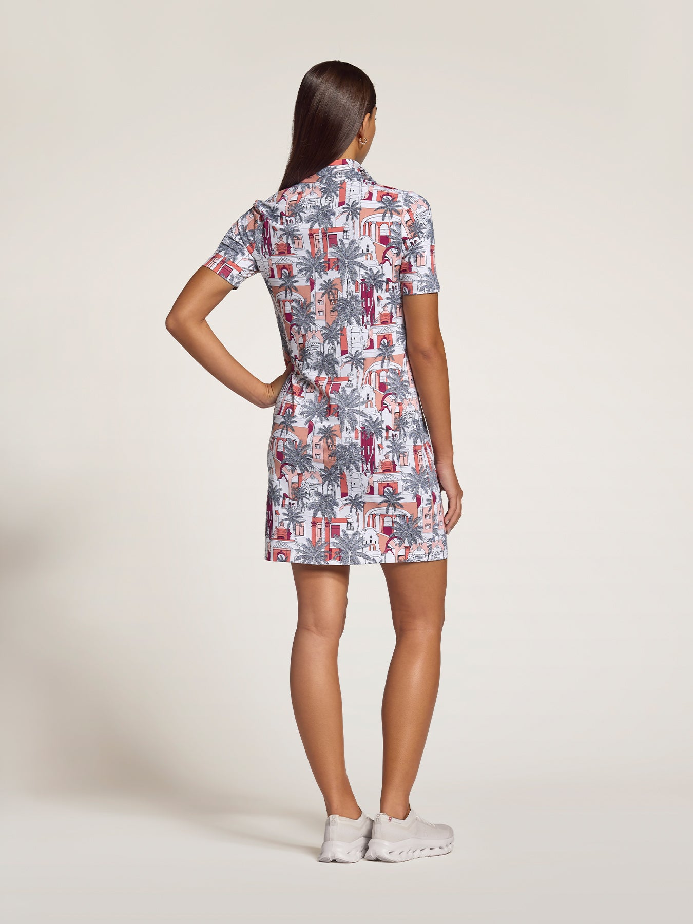 A woman is shown from the back wearing a short-sleeve collared shirt dress with a colorful tropical and geometric print, paired with white sneakers, against a plain light beige studio background.
