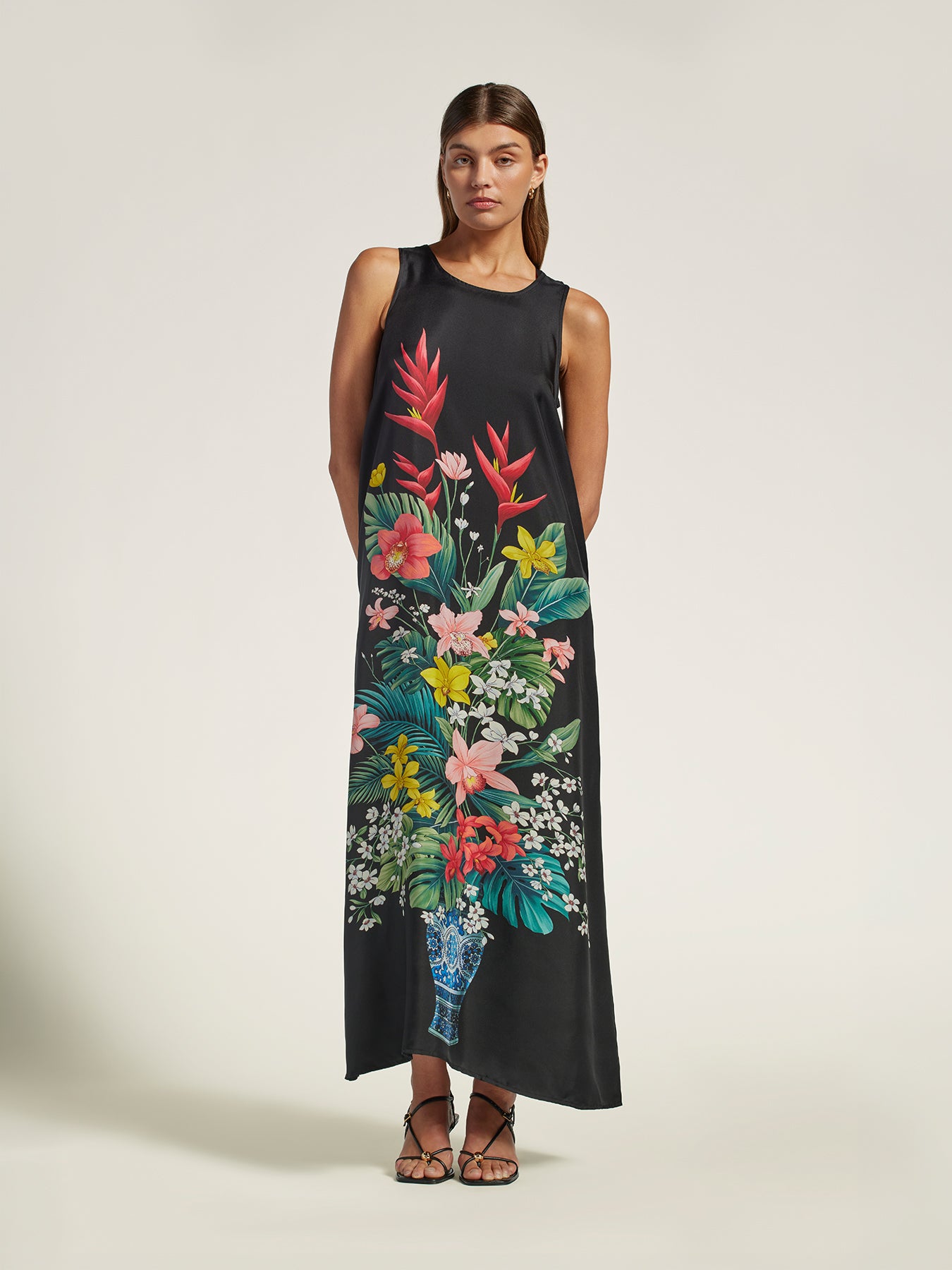 A woman modeling a sleeveless black maxi dress with a large, vibrant tropical floral print in the center, styled with brown strappy sandals against a plain light beige studio background.