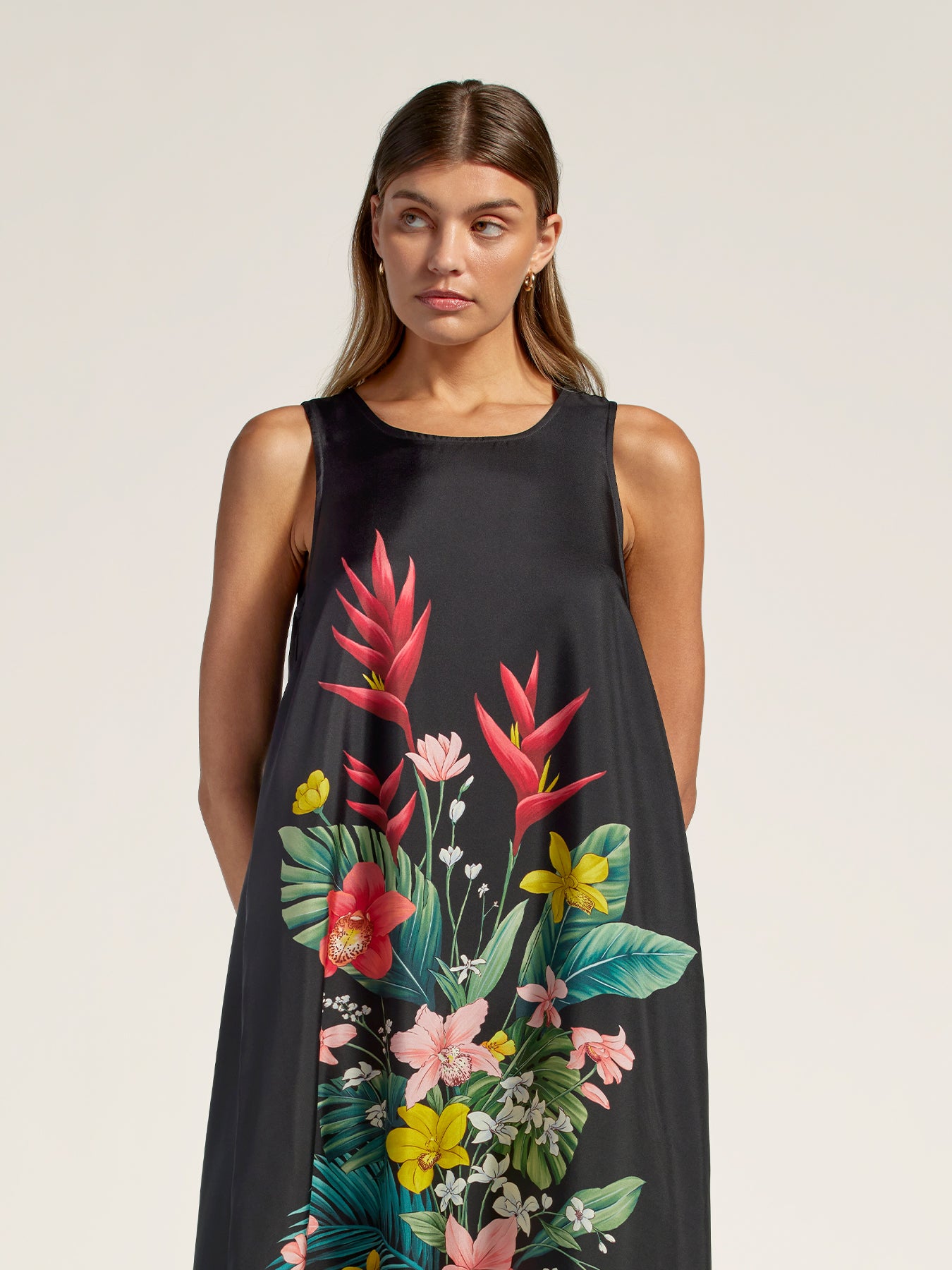 A woman modeling a sleeveless black dress with a vibrant tropical floral print featuring pink, red, yellow, and green flowers and leaves, set against a plain light beige background.