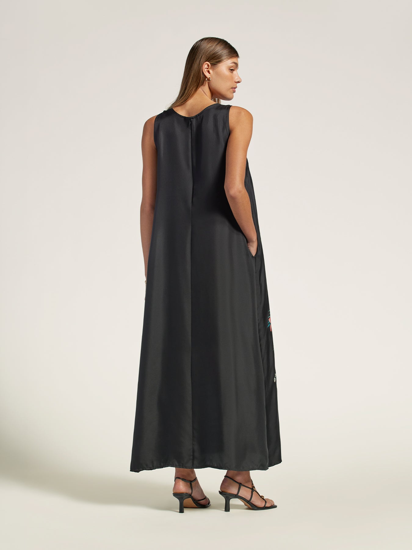 A woman modeling a sleeveless black maxi dress from the back, featuring a draped, open-back design and side pockets, set against a plain light beige studio background.