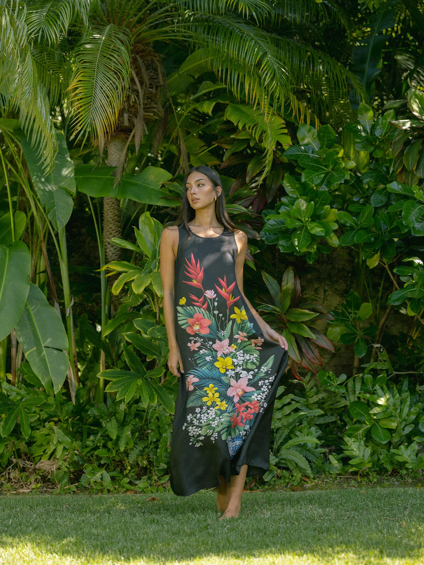 A woman wearing a sleeveless black maxi dress with a colorful tropical floral print stands in a lush green garden.