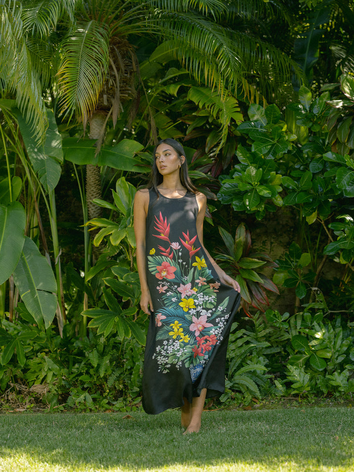 A woman wearing a sleeveless black maxi dress with a colorful tropical floral print stands in a lush green garden.