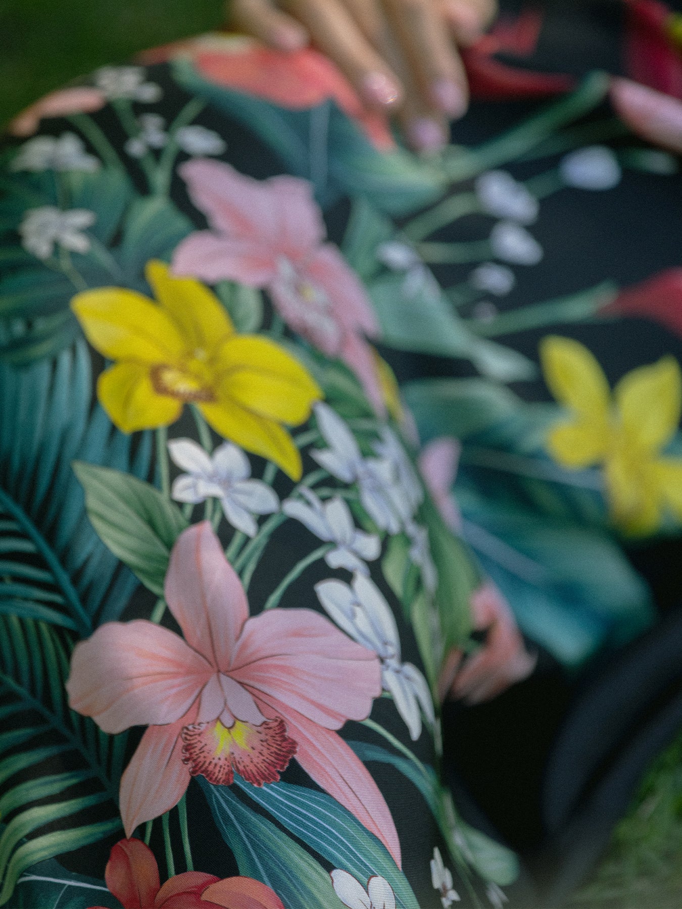 A close-up of a tropical floral print fabric featuring pink orchids, yellow flowers, and green leaves on a dark background, with a hand touching the material.
