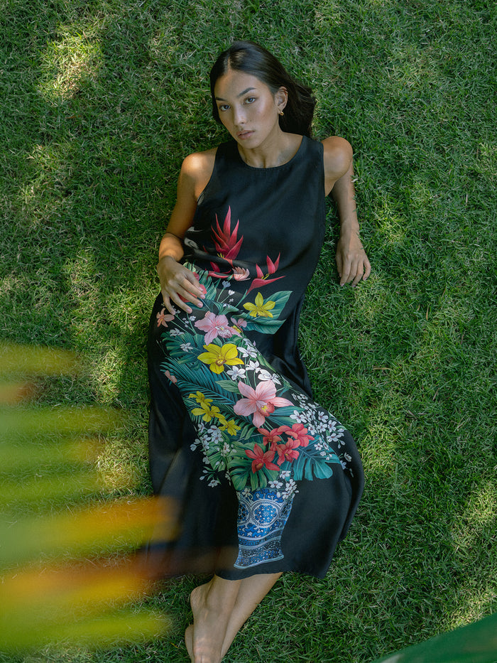 A woman lies on green grass wearing a sleeveless black maxi dress with a colorful tropical floral print. The dress features red, yellow, pink, and blue flowers with green leaves, and a blue and white pattern near the hem.