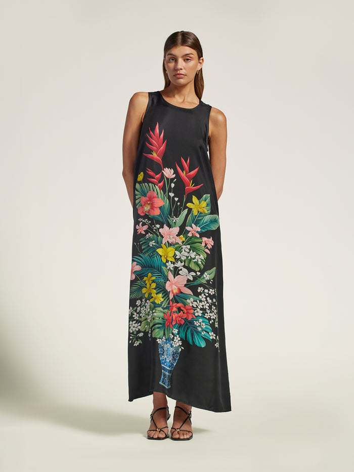 A woman modeling a sleeveless black maxi dress with a large, vibrant tropical floral print in the center, styled with brown strappy sandals against a plain light beige studio background.
