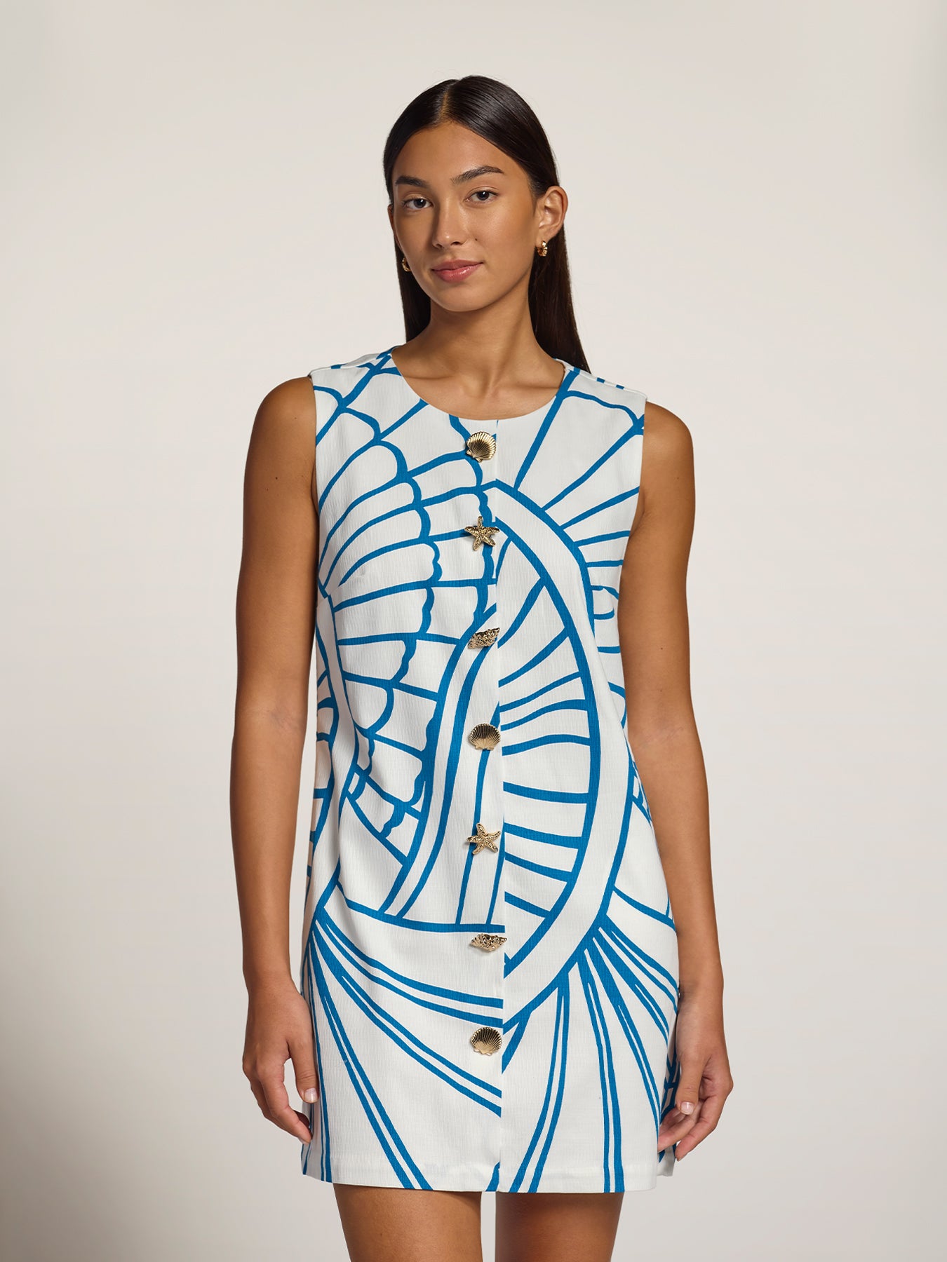 A woman modeling a sleeveless white dress with a bold blue abstract line pattern and a front button closure, photographed against a plain light beige studio background.